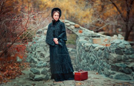 Beautiful woman in black long vintage dress and hood standing near stone wall. Art work of romantic woman with a valise. Pretty tenderness model dreaming and looking at camera. Woman traveling.