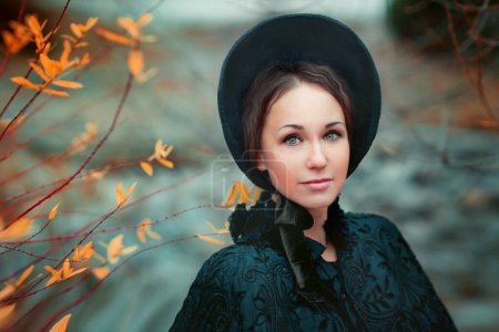 Close up portrait of Beautiful woman in black long vintage dress and hood standing near stone wall. Art work of romantic woman with a valise. Pretty tenderness model dreaming and looking at camera. Woman traveling.