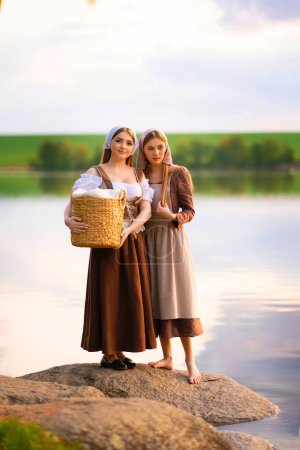 Pretty young two laundresses in medieval costume washing dresses near river .Beautiful girl working in countryside. Fairytale art work.