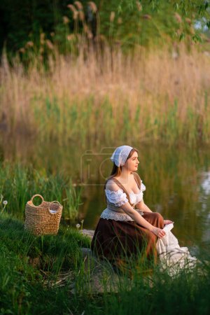 Hübsche junge Wäscherin in mittelalterlichem Kostüm steht am Fluss. Märchenhafte Kunstwerke.