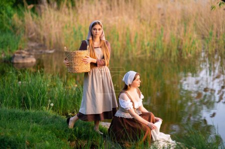 Pretty young two laundresses in medieval costume washing dresses near river .Beautiful girl working in countryside. Fairytale art work.