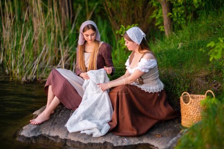 Téléchargez les photos : Jolies jeunes deux blanchisseries en costume médiéval robes de lavage près de la rivière.Belle fille qui travaille dans la campagne. Œuvre d'art conte de fées. - en image libre de droit