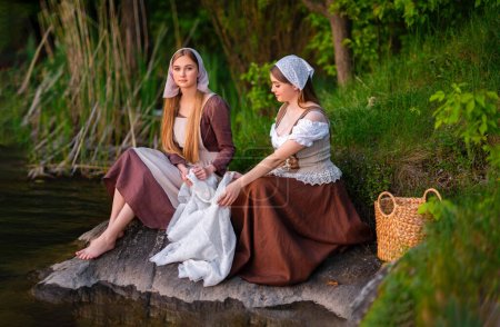 Pretty young two laundresses in medieval costume washing dresses near river. Beautiful girl working in countryside. Fairytale art work.