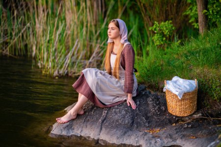 Bastante joven lavandera en traje medieval de pie cerca del río. Obra de arte de cuento.