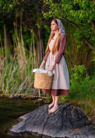 Pretty young laundress in medieval costume standing near river. Fairytale art work.