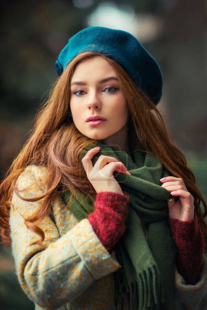 Foto de Retrato de una pelirroja hermosa niña en una bufanda verde y sombrero azul de pie sobre un fondo colorido. Obra de arte de la mujer romántica en otoño. Bonita modelo de ternura mirando a la cámara. - Imagen libre de derechos