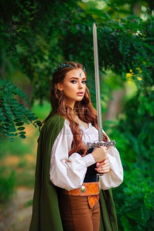 Téléchargez les photos : Belle princesse aux cheveux rouges en robe d'elfe médiévale et long manteau avec épée debout en pose guerrière sur fond de nature verte. Histoire de conte de fées sur le guerrier en diadème. Travail d'art chaud et coloré. - en image libre de droit