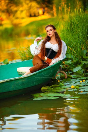Photo for Beautiful red haired princess in medieval elfin dress sitting in a boat on a river with green nature background. Fairy tale story and Warm colourful art work. - Royalty Free Image
