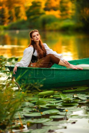 Téléchargez les photos : Belle princesse aux cheveux roux en robe d'elfe médiévale assise dans un bateau sur une rivière avec un fond vert nature. Conte de fées et oeuvre d'art chaude et colorée. - en image libre de droit