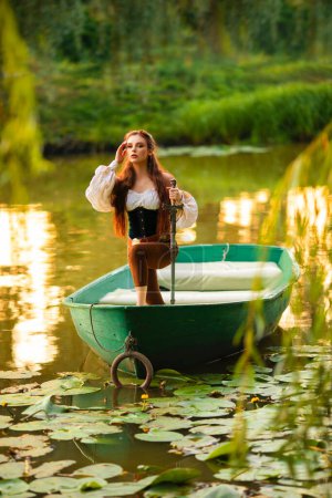Photo for Beautiful red haired princess in medieval elfin dress standing in a boat on a river with green nature background. Fairy tale story and Warm colourful art work. - Royalty Free Image