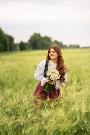 Photo for Beautiful red haired girl in  medieval dress standing in a wheat field with daisies . Fairy tale story about tenderness cute woman .Warm art work. - Royalty Free Image