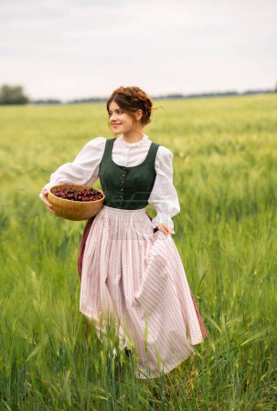 Beautiful red haired girl in  medieval dress standing in a wheat field with cherries . Fairy tale story about tenderness cute woman .Warm art work.