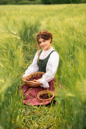 Photo for Beautiful red haired girl in  medieval dress standing in a wheat field with cherries . Fairy tale story about tenderness cute woman .Warm art work. - Royalty Free Image