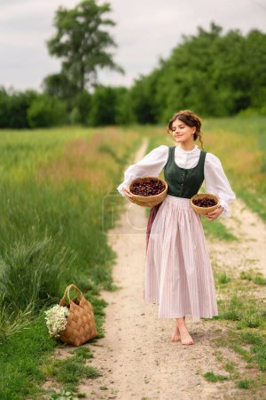 Téléchargez les photos : Belle fille aux cheveux roux en robe médiévale debout dans un champ de blé avec des cerises. Conte de fées histoire de tendresse femme mignonne .Travail d'art chaud. - en image libre de droit