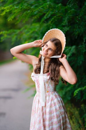 Téléchargez les photos : Jeune jolie fille blonde bouclée en chapeau de paille et robe vintage posant dans la nature avec un fond vert. Belle dame élégante dans le bonnet sur le travail d'art chaud coloré. - en image libre de droit