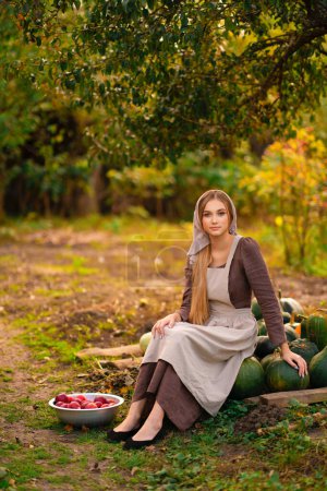 Téléchargez les photos : Belle jeune fille rurale en robe médiévale et bonnet posant sur un ranch. Jolie mannequin blonde en photo d'art de la campagne. - en image libre de droit