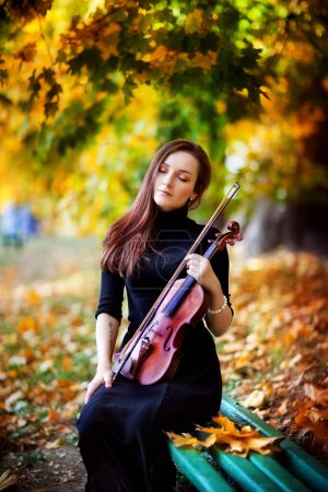 Foto de Retrato de una hermosa chica morena en vestido oscuro con violín. Violinista con los ojos cerrados en un parque otoñal. - Imagen libre de derechos