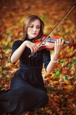 Photo for Portrait of a beautiful brunette girl in dark dress with violin. Violinist with sad eyes playing in autumn park. - Royalty Free Image