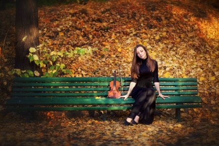 Photo for Portrait of a beautiful brunette girl in dark dress with violin. Violinist with sad eyes on yellow background in a autumn park. - Royalty Free Image