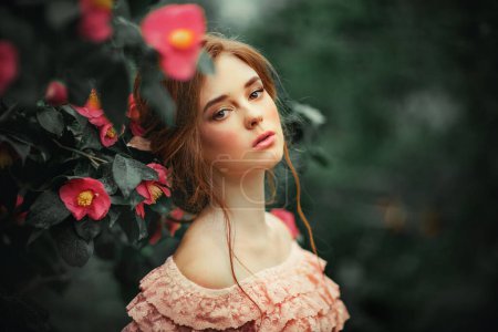 Téléchargez les photos : Gros plan portrait d'une belle fille aux cheveux roux dans une robe vintage rose debout près de fleurs colorées. Oeuvre d'art de femme romantique. Jolie modèle de tendresse regardant la caméra. - en image libre de droit