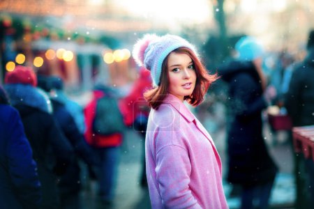 Photo for Portrait of a beautiful young model in a knitting cap standing on a christmas background with bokeh lights. Magic warm new year photo.Cozy background of winter fair. Happy woman. - Royalty Free Image
