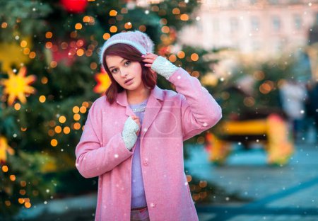 Téléchargez les photos : Portrait d'un beau jeune modèle dans une casquette en tricot debout sur un fond de Noël avec des lumières bokeh. Magie chaude nouvelle année photo.Cozy fond de foire d'hiver. Femme heureuse. - en image libre de droit