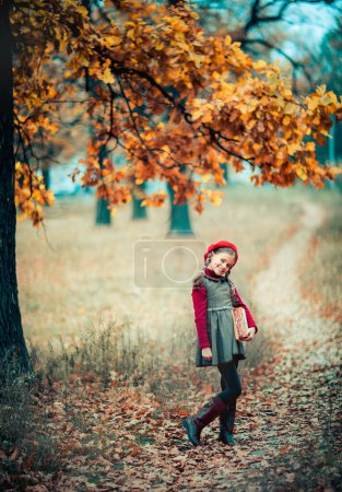 Photo for Autumn portrait of a beautiful young schoolgirl in red beret and braid hairs with books . Tenderness positive child with bright smile enjoying nature in park. - Royalty Free Image