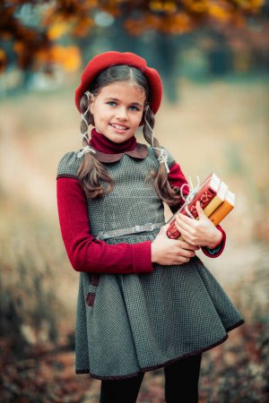 Photo for Autumn portrait of a beautiful young schoolgirl in red beret and braid hairs with books . Tenderness positive child with bright smile enjoying nature in park. - Royalty Free Image