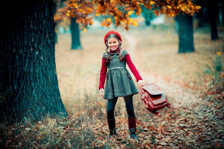 Photo for Autumn portrait of a beautiful young schoolgirl in red beret and braid hairs with school bag . Tenderness positive child with bright smile enjoying nature in park. - Royalty Free Image