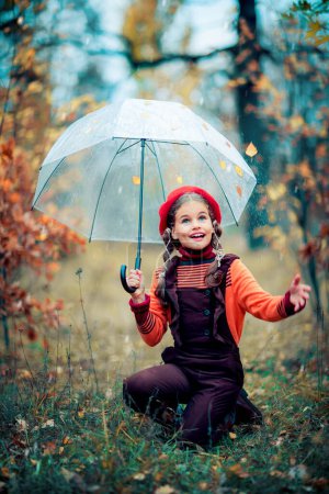 Photo for Autumn portrait of a beautiful young schoolgirl in red beret and braid hairs with umbrella and leaves on it. Tenderness positive child with bright smile enjoying nature in park. - Royalty Free Image