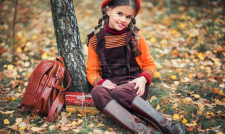 Téléchargez les photos : Portrait d'automne d'une belle jeune écolière en béret rouge et poils tressés avec livres. Sensibilité enfant positif avec sourire lumineux profiter de la nature dans le parc. - en image libre de droit
