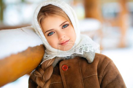 Foto de Retrato de invierno de una hermosa joven con el pelo largo trenzado, ojos azules y bufanda de encaje de punto de pie sobre un fondo de madera y copos de nieve. - Imagen libre de derechos