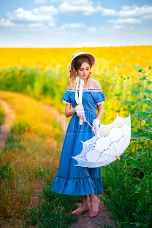 Téléchargez les photos : Jolie jeune fille bouclée en chapeau de paille et robe jaune vintage debout près des fleurs blanches. Dame élégante marchant dans la campagne.Oeuvre d'art de modèle de tendresse romantique. - en image libre de droit