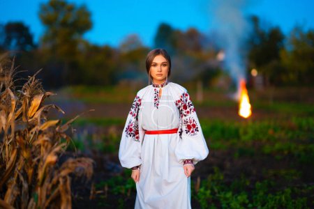 Foto de Hermosa chica rubia en vestido tradicional ucraniano en otoño maíz field.Bonfire en el fondo. - Imagen libre de derechos