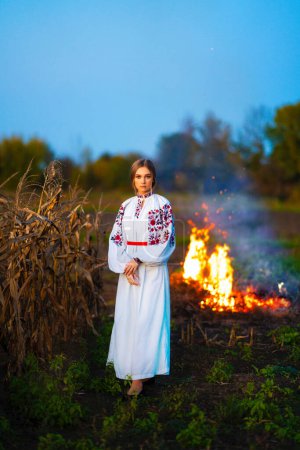 Photo for Beautiful blonde girl in Ukrainian traditional dress in autumn corn field.Bonfire on background. - Royalty Free Image
