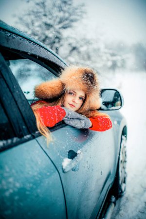 Foto de Hermosa chica rubia de pelo largo con ojos azules en suéter rojo y gorra de piel de pie cerca del coche en las nevadas de invierno. - Imagen libre de derechos