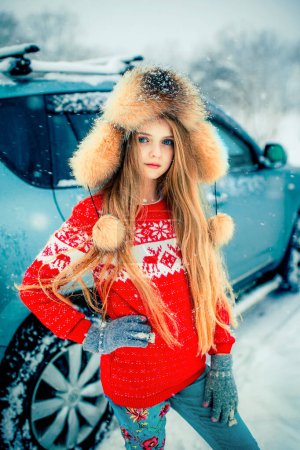 Foto de Hermosa chica rubia de pelo largo con ojos azules en suéter rojo y gorra de piel de pie cerca del coche en las nevadas de invierno. - Imagen libre de derechos