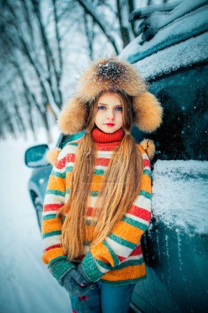 Foto de Hermosa chica rubia de pelo largo con ojos azules en suéter y gorra de piel de pie cerca del coche en las nevadas de invierno. - Imagen libre de derechos
