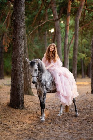 Téléchargez les photos : Belle fille aux cheveux longs en robe longue glamour posant avec cheval gris dans la forêt de pins. Travail d'art de conte de fées chaud - en image libre de droit