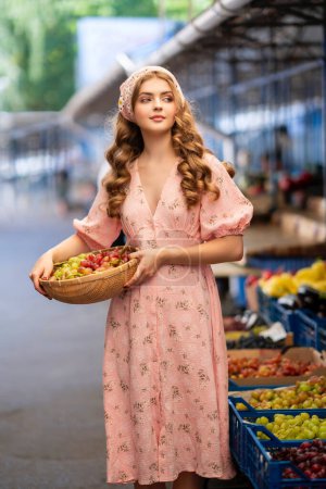 Hermosa chica rubia rizada en bufanda floral de punto y vestido rosa de pie en el mercado con diferentes frutas. Joven adolescente como vendedora con canasta de uvas en sus manos.