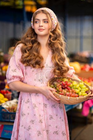 Foto de Hermosa chica rubia rizada en bufanda floral de punto y vestido rosa de pie en el mercado con diferentes frutas. Joven adolescente como vendedora con canasta de uvas en sus manos. - Imagen libre de derechos