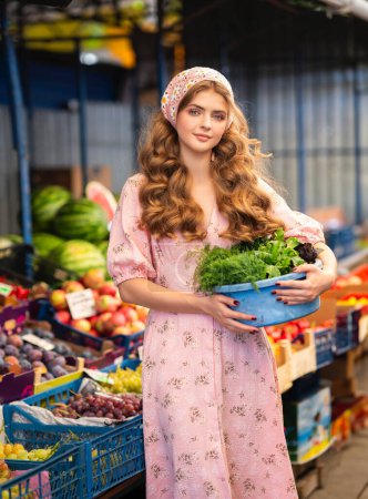 Photo for Beautiful blonde curly girl in knitted floral scarf and pink dress standing on market with different fruits. Young teenager as a seller with basket of grapes in her hands. - Royalty Free Image