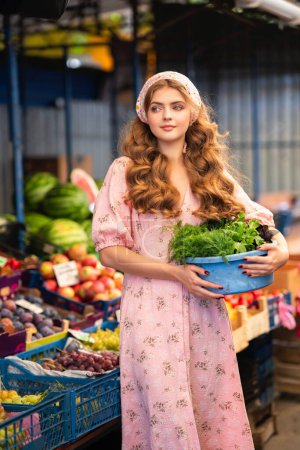Foto de Hermosa chica rubia rizada en bufanda floral de punto y vestido rosa de pie en el mercado con sandías. Joven adolescente vendiendo frutas. - Imagen libre de derechos