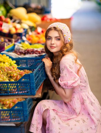 Foto de Hermosa chica rubia rizada en bufanda floral de punto y vestido rosa de pie en el mercado con diferentes frutas. Joven adolescente vendiendo verduras. - Imagen libre de derechos