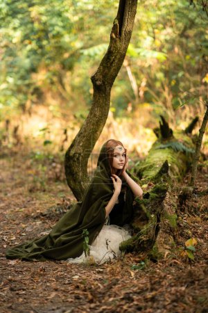 Photo for Portrait of  a beautiful red haired girl in green medieval dress near moss tree. Fairy tale story about elven.Amazing model posing in forest .Warm art work - Royalty Free Image