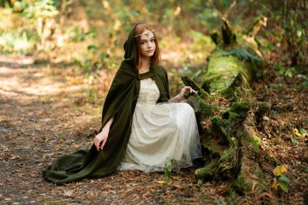 Photo for Portrait of  a beautiful red haired girl in green medieval dress near moss tree. Fairy tale story about elven.Amazing model posing in forest .Warm art work - Royalty Free Image