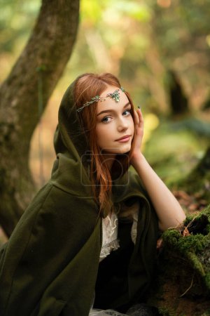 Téléchargez les photos : Portrait d'une belle fille aux cheveux roux en robe médiévale verte près d'un mousse. Histoire de conte de fées sur elven.Amazing modèle posant dans la forêt.Travail d'art chaud - en image libre de droit