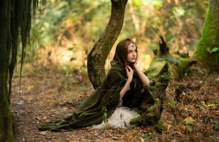 Téléchargez les photos : Portrait d'une belle fille aux cheveux roux en robe médiévale verte près d'un mousse. Histoire de conte de fées sur elven.Amazing modèle posant dans la forêt.Travail d'art chaud - en image libre de droit
