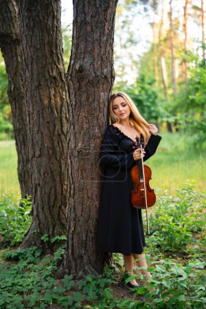 Photo for Beautiful blonde girl in black dress holding a violin in green park. Art work. Sad lady dreaming. - Royalty Free Image
