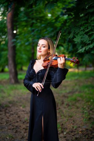 Foto de Hermosa chica rubia en vestido negro tocando el violín en el parque verde. Trabajo artístico. Triste dama soñando. - Imagen libre de derechos